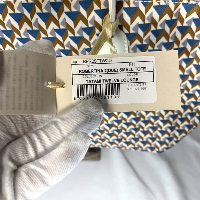Ръкавицата държи етикета на продукта "ROBERTINA 2 (DUE) SMALL TOTE" върху тъканта с татами с геометричен модел в синьо, кафяво и бяло, което е характерният стил на малката колекция чанта на ROBERTA PIERI.