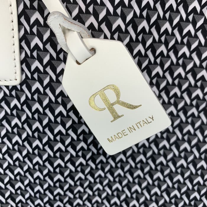 Бял кожен етикет ROBERTA PIERI с буквите "R" в злато и думите "ПРОИЗВЕДЕНО В ИТАЛИЯ" виси на фона на черно-бяла геометрична материя.